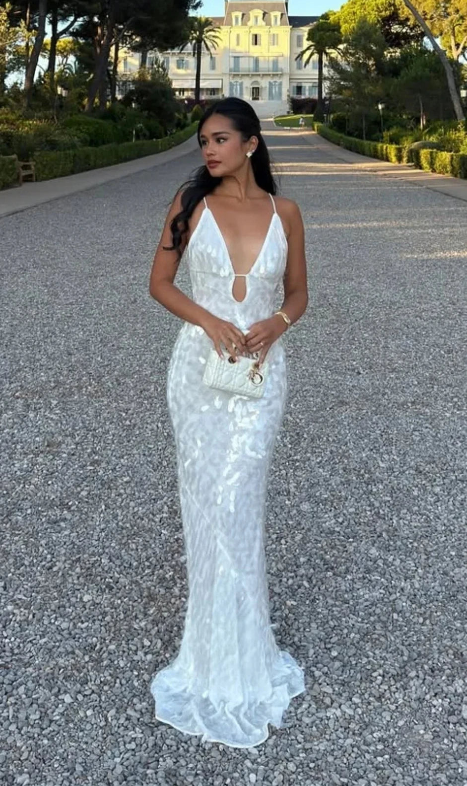 A woman wearing a white lace mermaid-style evening gown stands on a gravel path outdoors, with trees and a building in the background.