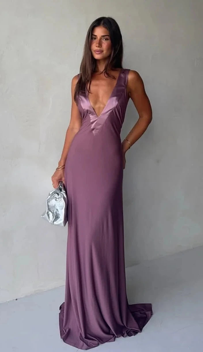Woman in a long purple dress holding a silver clutch against a plain background