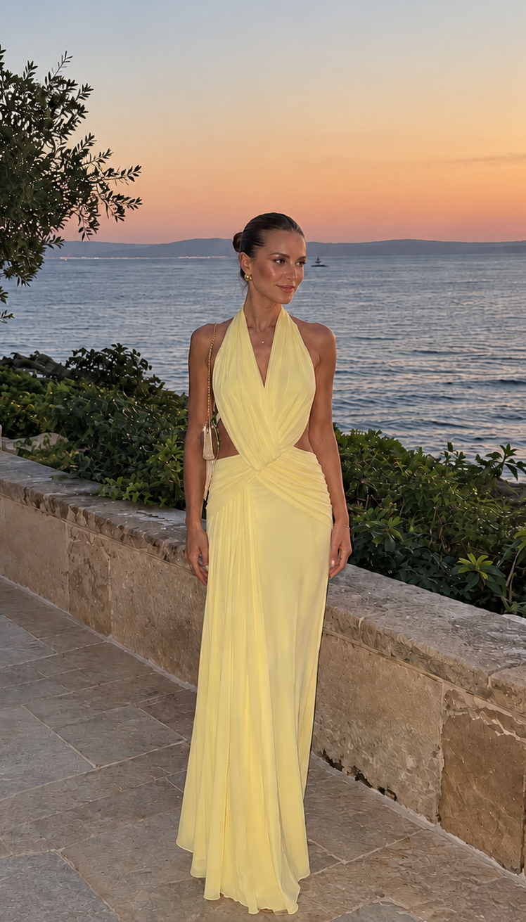 A woman wearing a pale yellow halter-neck maxi dress with a side cutout, standing on a stone terrace overlooking the sea at sunset.